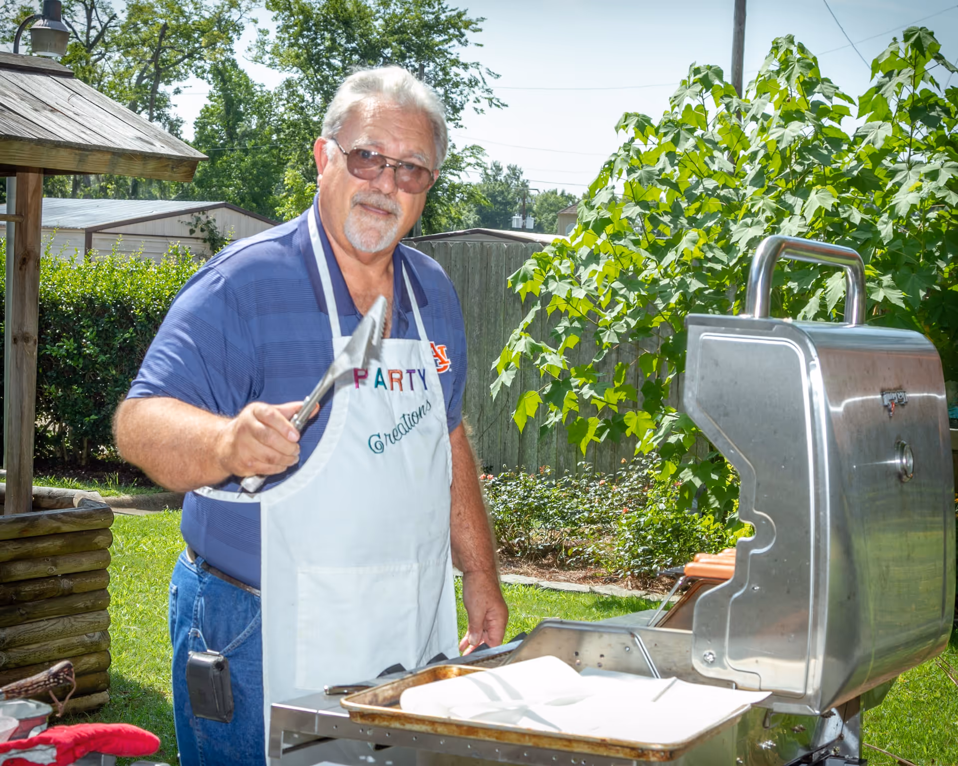 An older man wearing sunglasses and a white apron that says 'Party Creations' is grilling outdoors. He is holding a pair of tongs and standing next to a stainless steel grill with hot dogs cooking on it. The background shows green bushes, trees, and a wooden fence under a clear sky.