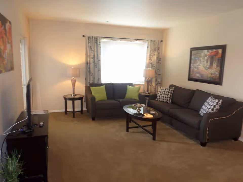 A cozy living room with two dark gray sofas adorned with green and patterned pillows, a glass-top coffee table in the center, two side tables each with a lamp, a window with floral curtains, a TV on a stand to the left, and framed artwork on the walls.