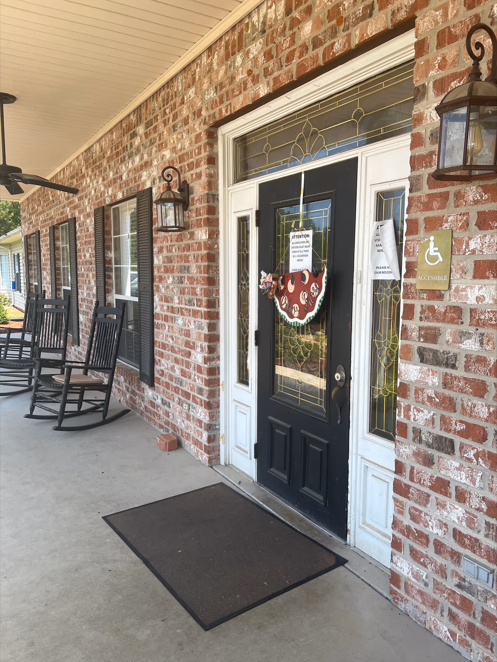 Brick front porch showing a black decorative entrance door with sidelights, rocking chairs, and an accessible sign.