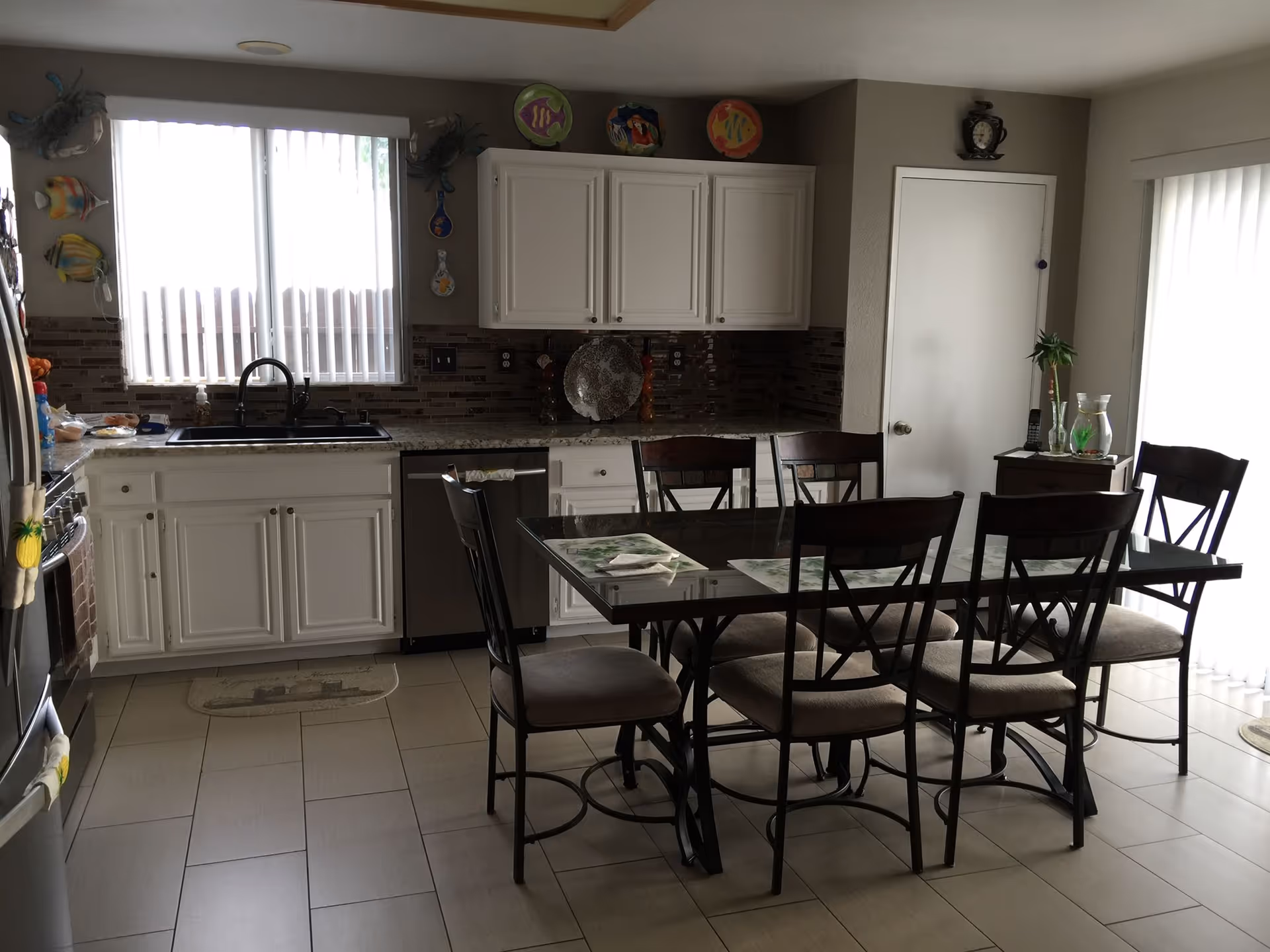 A kitchen and dining area with white cabinets, a black sink, and a glass-top dining table with six cushioned chairs. The kitchen has a window with vertical blinds, decorative fish wall hangings, and a tiled backsplash. There is a small side table with plants and glass pitchers near a sliding glass door with vertical blinds.