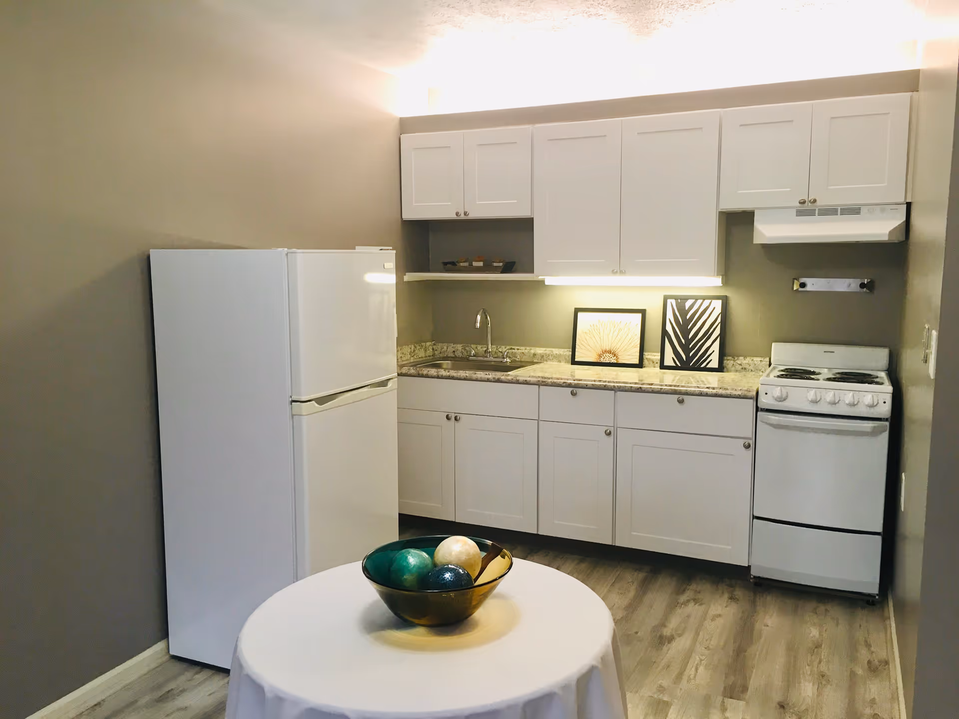 A small kitchen area with white cabinets, a white refrigerator, a white stove with an overhead vent, and a countertop with a sink. Two framed pictures are placed on the countertop. In the foreground, there is a round table covered with a white tablecloth and a decorative bowl containing three large decorative balls.