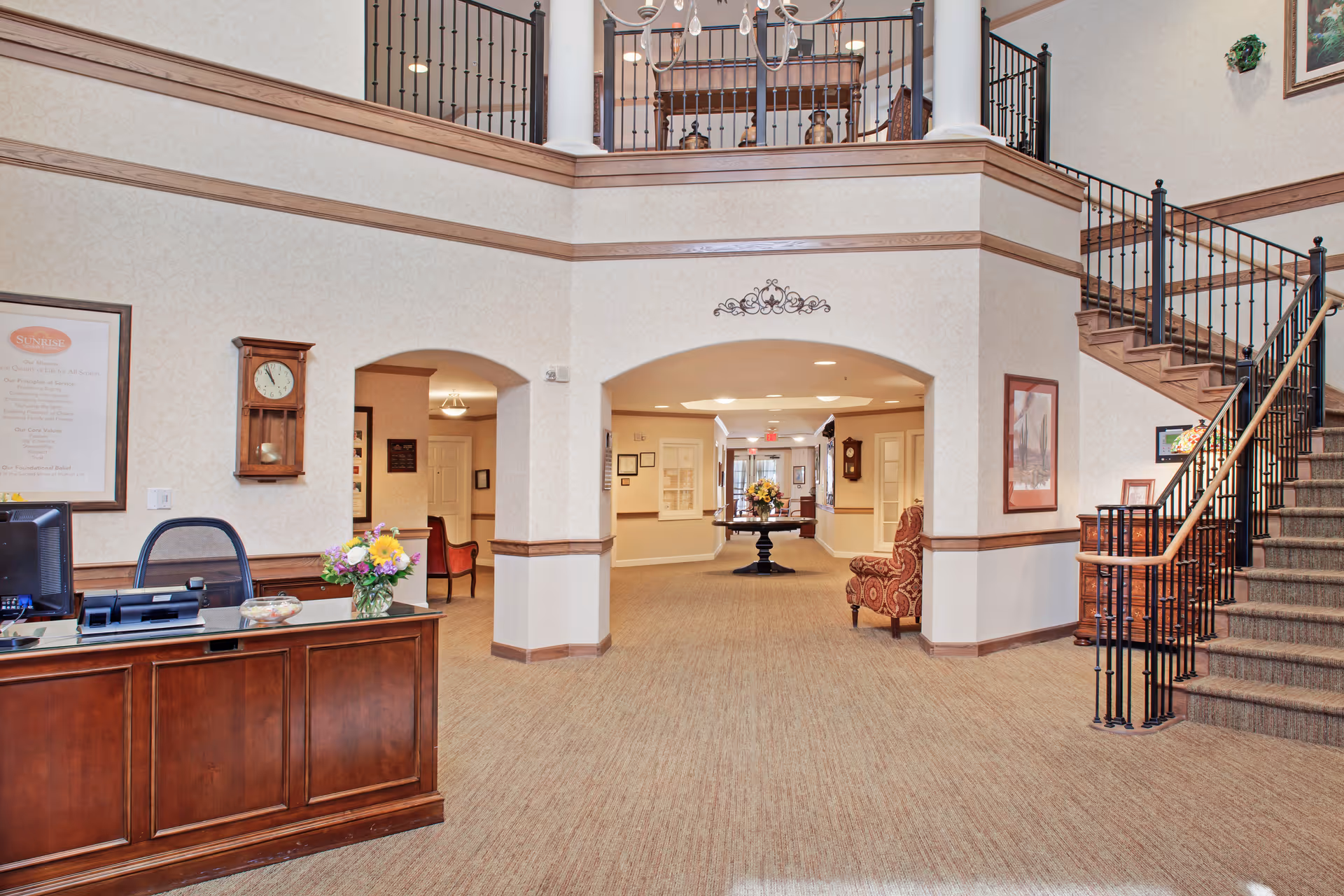 Spacious two-story assisted-living lobby with a reception desk, central table of flowers, and a staircase to an upper level.
