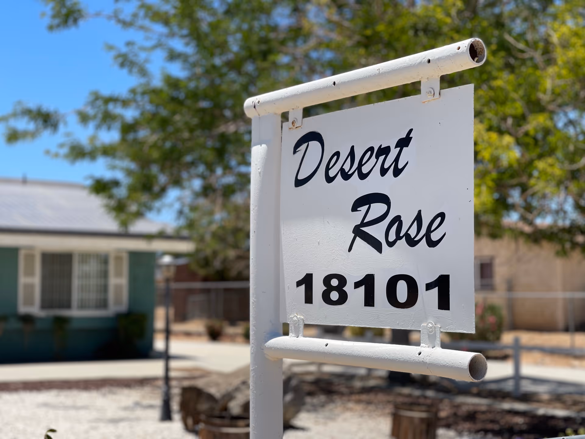 A white sign with black text reading 'Desert Rose 18101' mounted on a white metal frame outdoors, with a building, trees, and a clear blue sky in the background.