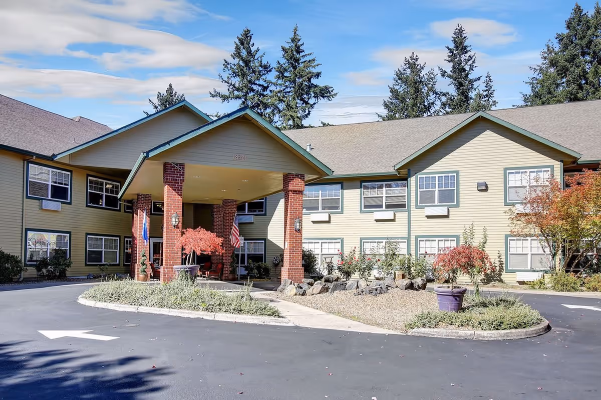 Exterior view of a senior living facility building with a covered entrance supported by brick columns. The building is two stories high with multiple windows and beige siding. There are small landscaped areas with plants and trees near the entrance, and a paved driveway with a white directional arrow.