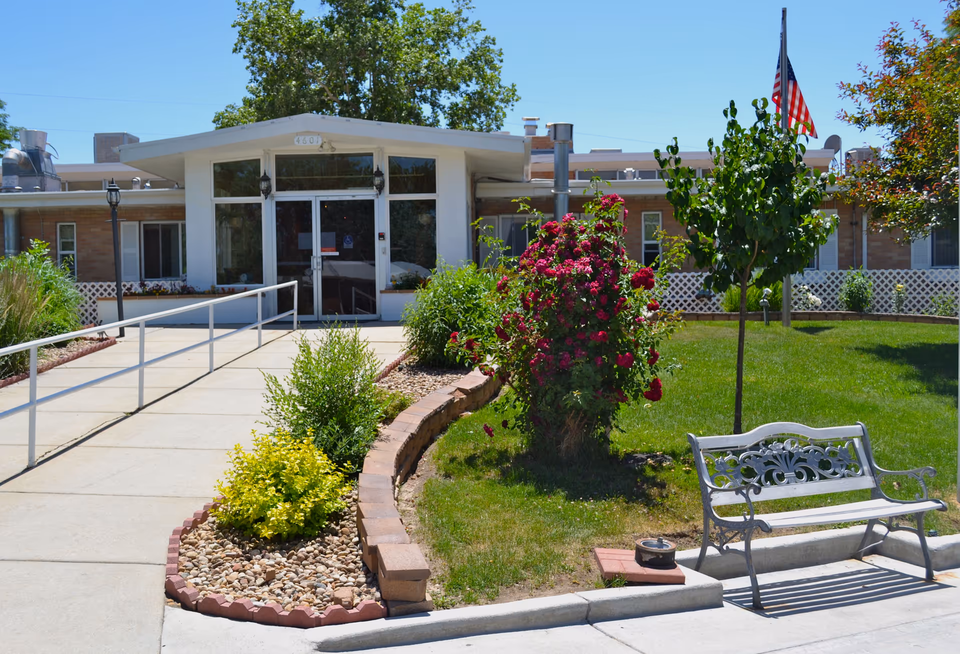Front entrance of a single-story building with a wheelchair ramp, landscaped flower beds, a bench, and an American flag.
