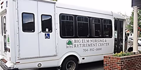 A white shuttle bus parked outside a facility displaying the 'Big Elm Nursing & Retirement Center' name and phone number beside a brick planter.