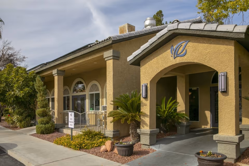 Exterior view of Villa Court Assisted Living facility showing a beige stucco building with arched entryways, decorative plants, and a sidewalk leading to the entrance under a partly cloudy sky.
