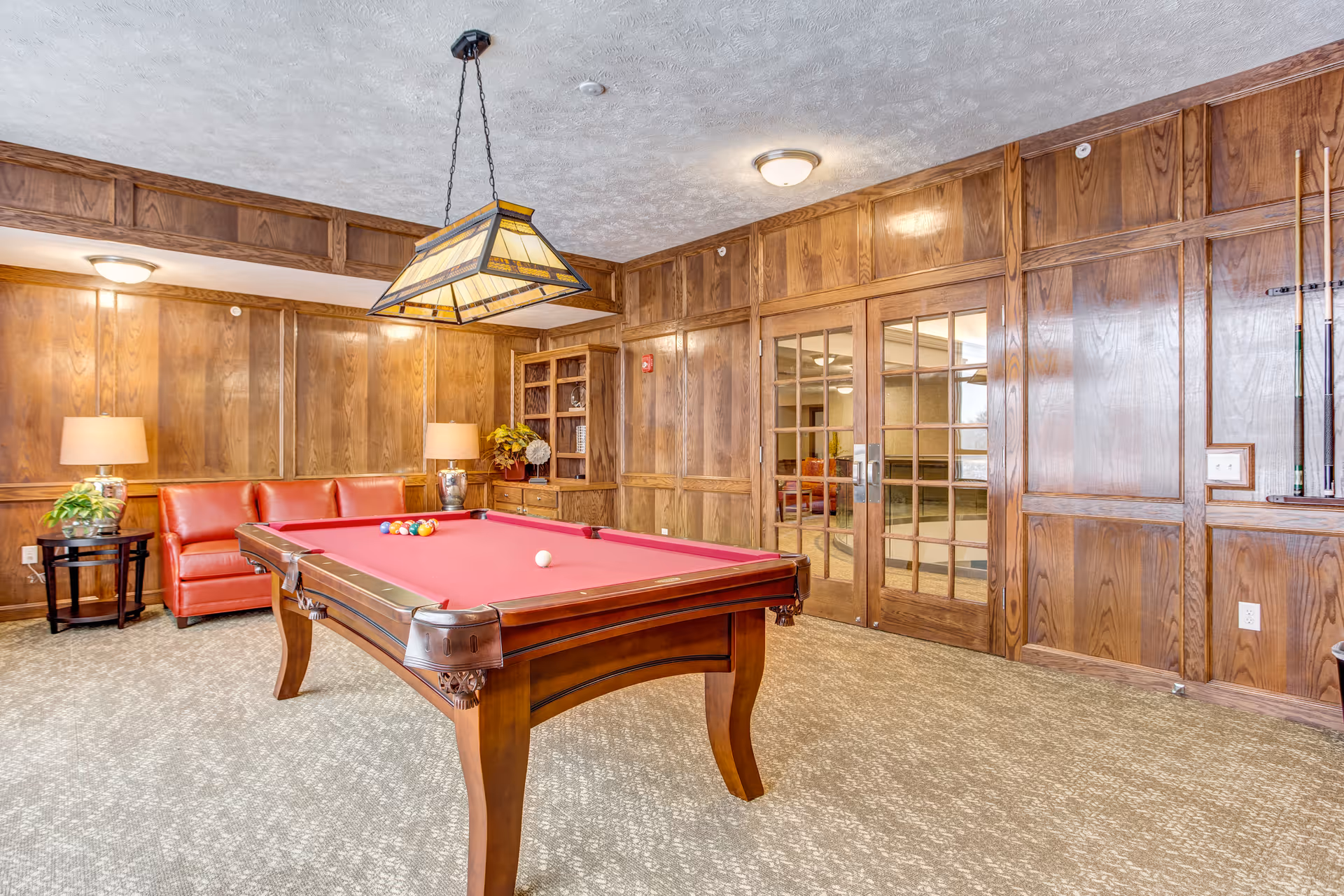 Wood-paneled recreation room featuring a red-felt pool table, red sofa, lamps, and glass double doors.