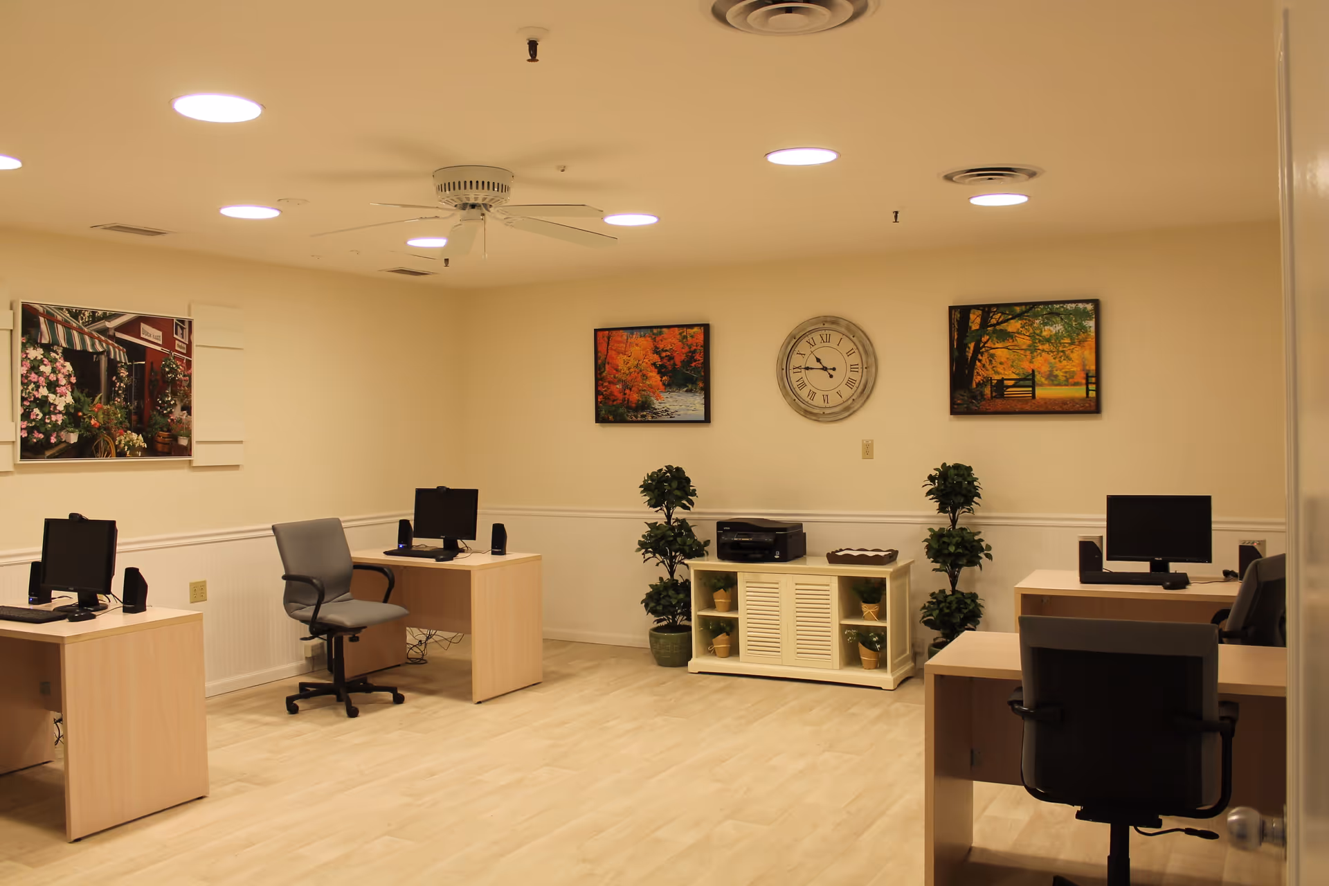 A well-lit office room with four desks, each equipped with a computer and a chair. The room has light-colored wooden flooring and cream-colored walls adorned with three framed pictures and a large round clock. There are two potted plants flanking a white cabinet with a printer on top.