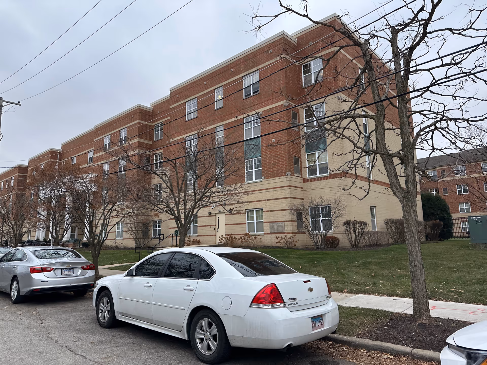 Exterior view of a multi-story brick residential building with several windows, leafless trees, and parked cars along the street in front. The sky is overcast.