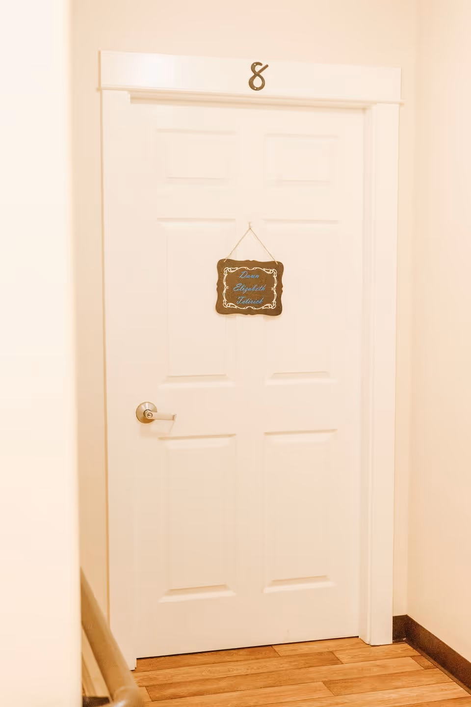 A closed white door with a silver handle in a hallway with light-colored walls and wooden flooring. The door has a small hanging sign with decorative edges and blue cursive text. Above the door frame is a metallic number 8.