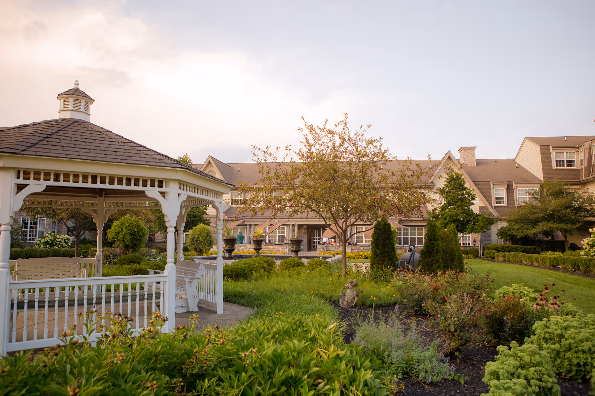 A peaceful outdoor garden area at The Manor at York Town featuring a white gazebo with a bench swing, surrounded by lush greenery, flowering plants, and trees, with a large residential building in the background under a partly cloudy sky.