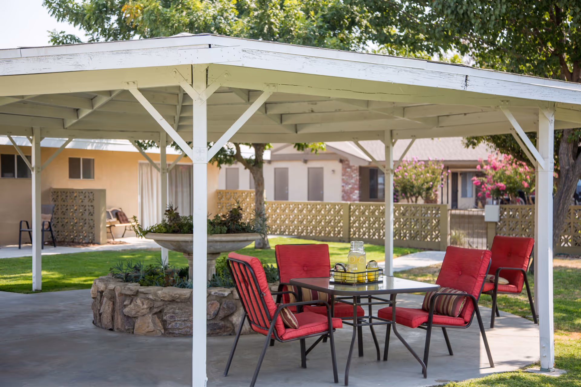 Outdoor covered seating area with a table and four red cushioned chairs around it. A glass pitcher with lemon water and glasses is on the table. The area is surrounded by green grass, trees, and residential buildings in the background.