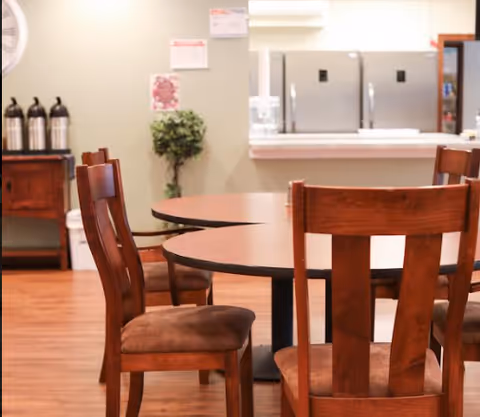Interior view of a dining area with wooden chairs and round tables. In the background, there is a counter with two refrigerators and a water dispenser. A small green plant is placed near the wall, and some notices are pinned on the wall above the plant.