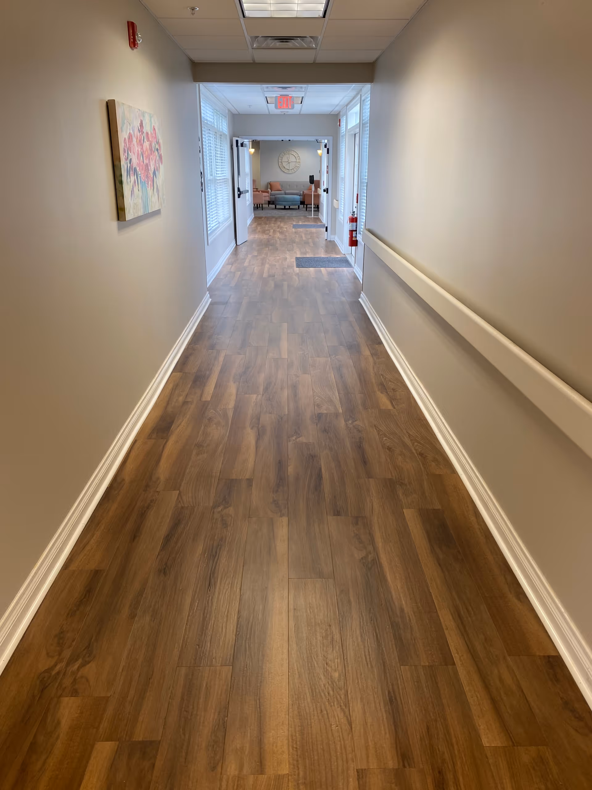 A long, clean hallway with wood-patterned flooring and beige walls. There is a handrail on the right wall and a floral painting on the left wall. At the end of the hallway, double doors open into a room with seating and a large wall clock.