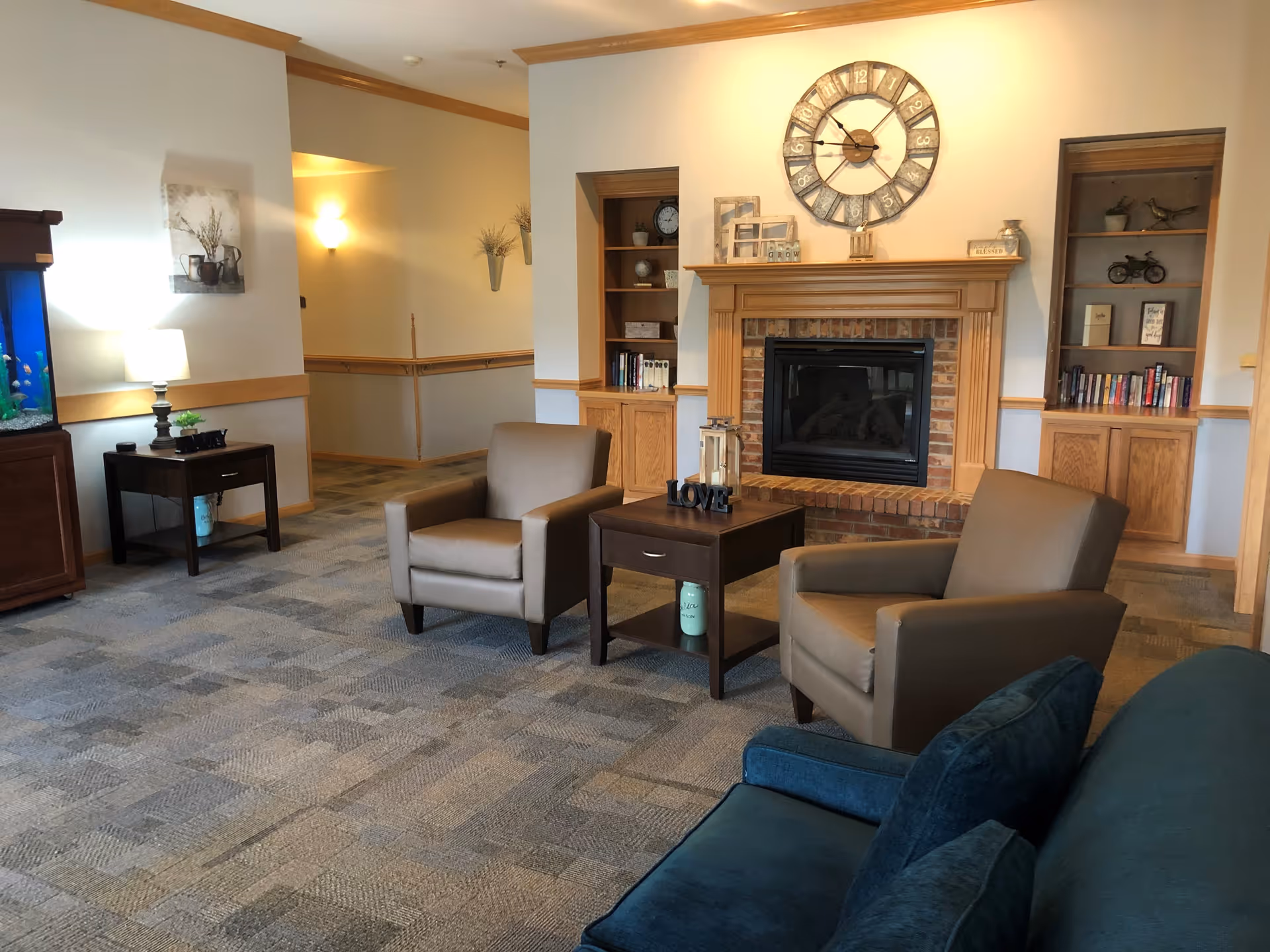 A cozy living room area with two beige armchairs and a dark wooden side table between them. The side table has decorative items including a lantern and a 'LOVE' sign. Behind the chairs is a fireplace with a large rustic clock above it and built-in wooden shelves on either side filled with books and decor. To the left, there is a small table with a lamp and a fish tank on a wooden stand. The room has carpeted flooring and soft lighting.