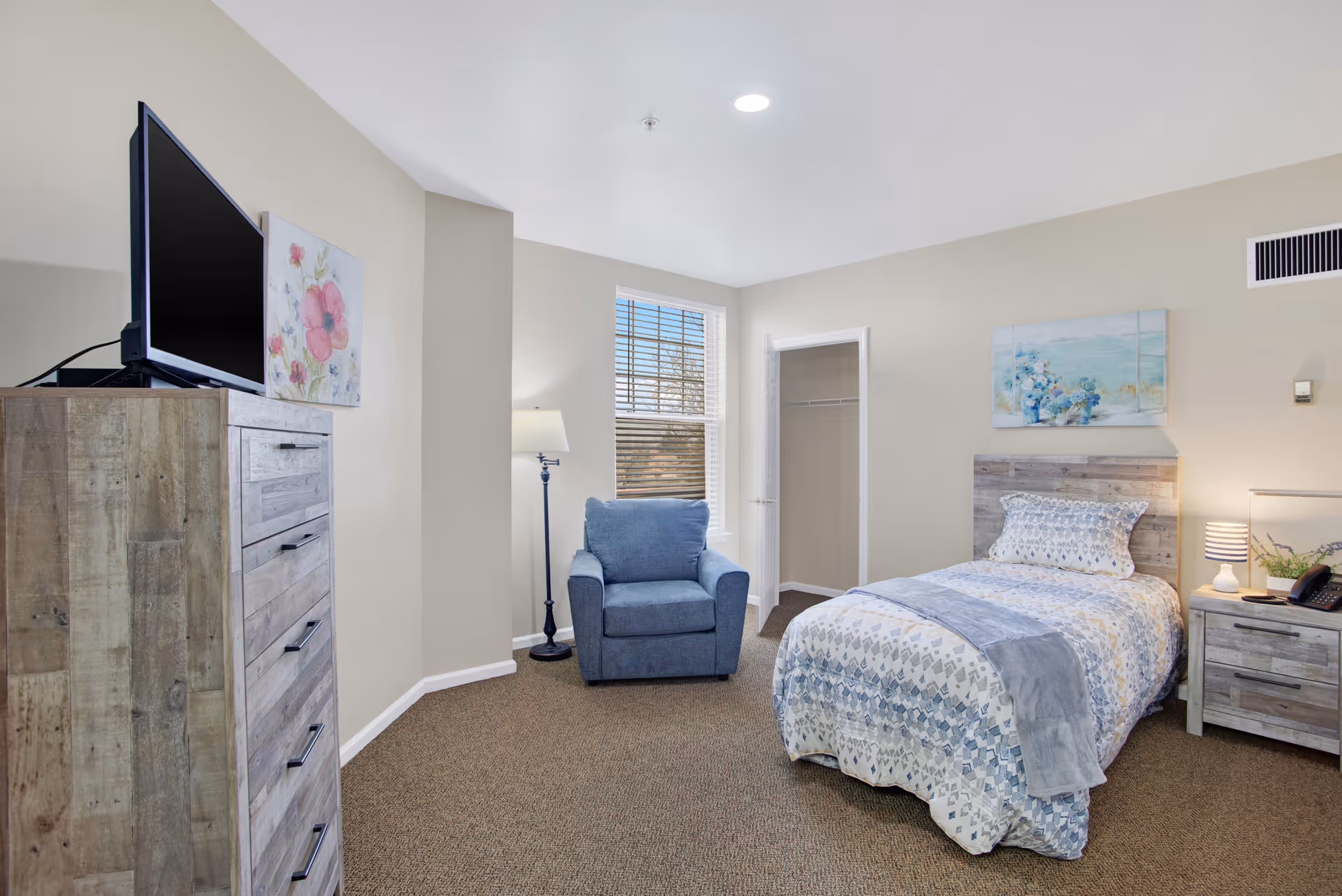 A cozy bedroom in a senior living facility featuring a single bed with patterned bedding and a gray throw blanket, a wooden nightstand with a lamp and a phone, a blue upholstered armchair next to a window with blinds, a tall wooden dresser with a flat-screen TV on top, and light-colored walls adorned with floral artwork.