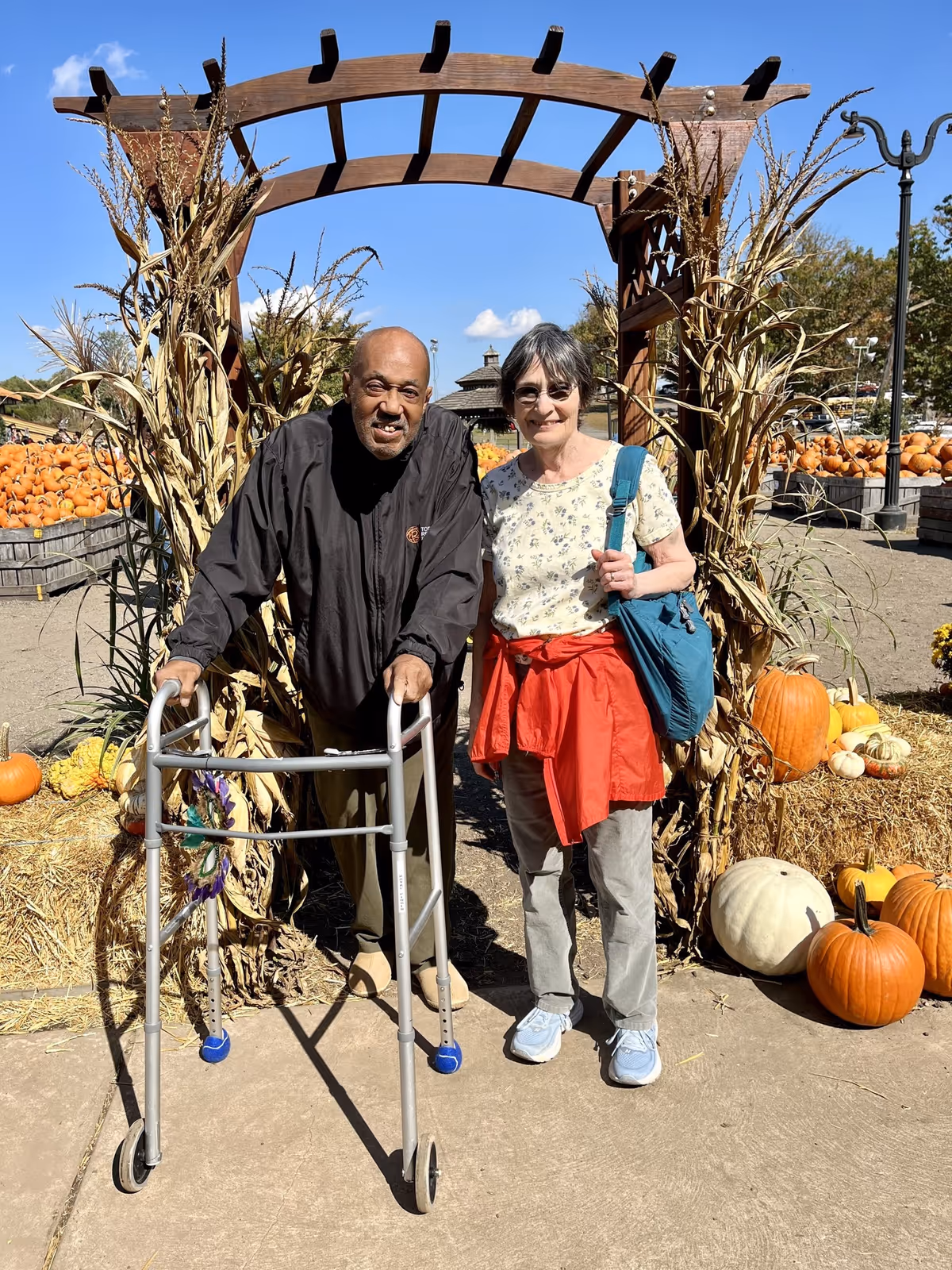 An elderly man using a walker and an elderly woman standing beside him under a wooden arch decorated with dried cornstalks. They are outdoors at a pumpkin patch with many pumpkins and hay bales around them under a clear blue sky.