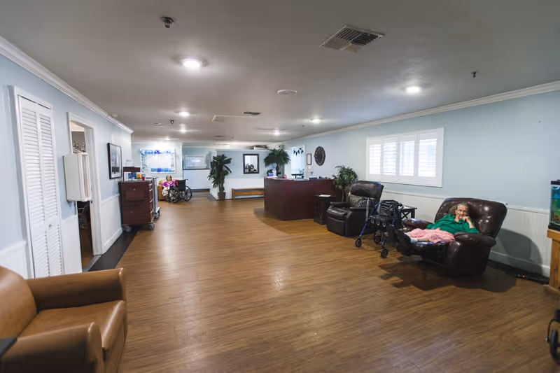 A spacious senior living facility common area with light blue walls and wood flooring. There are comfortable leather chairs, a reception desk, potted plants, and two elderly women, one sitting in a wheelchair and another reclining in a leather chair with a walker nearby. The room is well-lit with ceiling lights and has a window with white shutters.