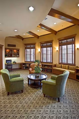 Cozy senior living lounge with green armchairs around a round table and large windows.