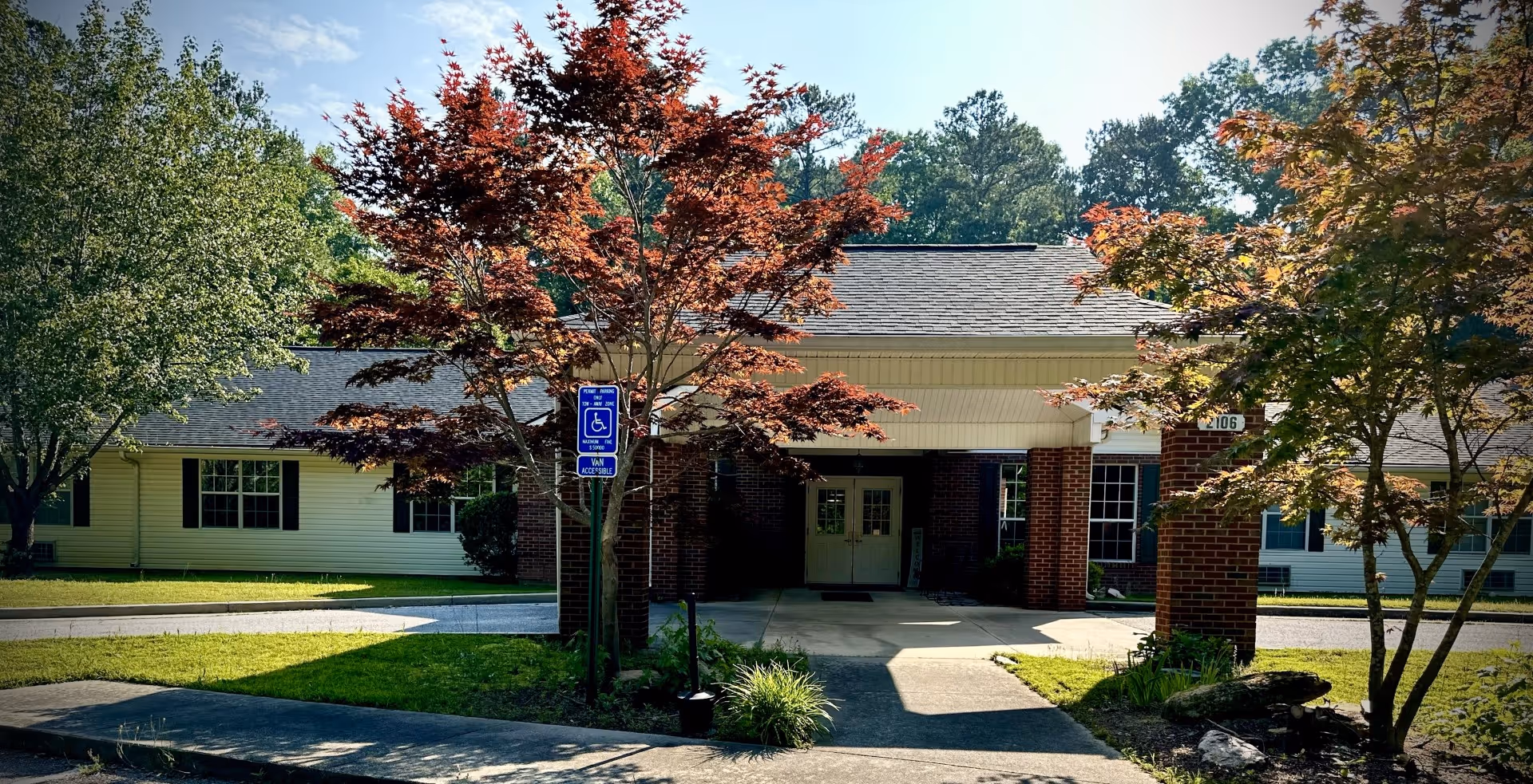 Front entrance of a single-story building with a covered portico, brick columns, landscaped trees, and a visible handicap parking sign.