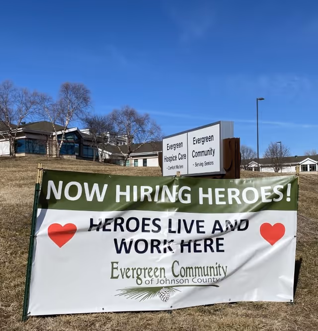 Outdoor scene showing a large banner in front of a building. The banner reads 'NOW HIRING HEROES! HEROES LIVE AND WORK HERE Evergreen Community of Johnson County' with two red heart icons. Behind the banner, there is a sign for Evergreen Hospice Care and Evergreen Community - Serving Seniors. The sky is clear and blue, and there are leafless trees and grass around the area.
