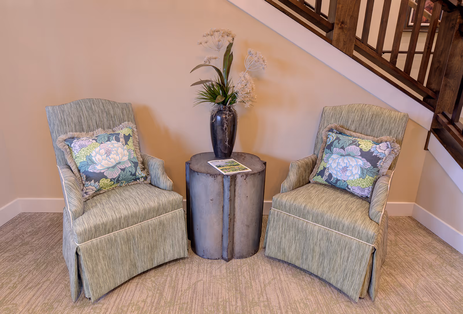 Two green upholstered armchairs with floral cushions flank a cylindrical metal side table holding a vase and a brochure beneath a wooden staircase.