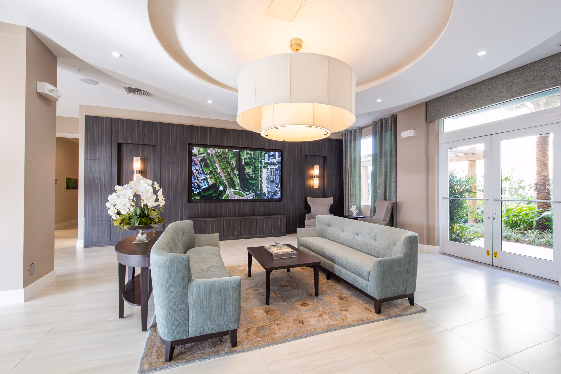A modern and bright lounge area with two light green sofas facing each other across a dark wooden coffee table on a patterned rug. Behind one sofa is a round side table with a white orchid plant. The back wall features a large flat-screen TV displaying an aerial view of a park, flanked by two wall sconces. Large windows and glass doors on the right side let in natural light and offer a view of greenery outside. The ceiling has a large circular recessed light fixture.