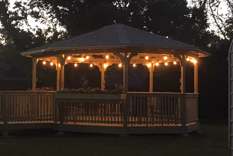 A wooden gazebo illuminated by string lights at dusk, surrounded by trees and grass.