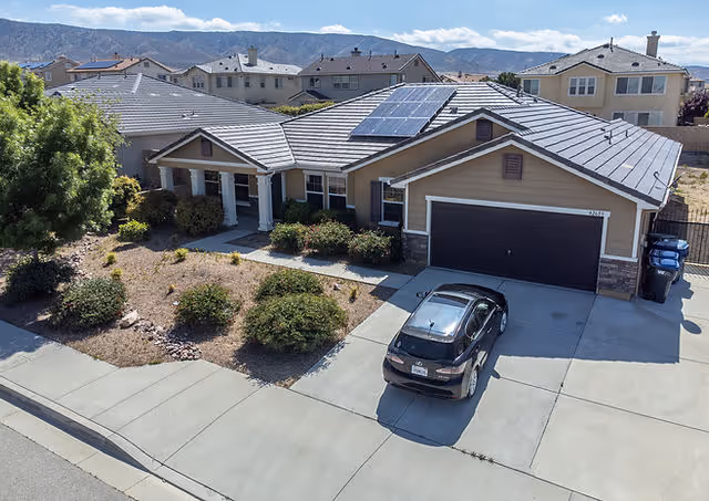 A single-story residential house with a gray tiled roof featuring solar panels, a two-car garage with a dark door, a black car parked in the driveway, and a landscaped front yard with bushes and a tree. The house is situated in a suburban neighborhood with other similar houses and mountains visible in the background under a partly cloudy sky.