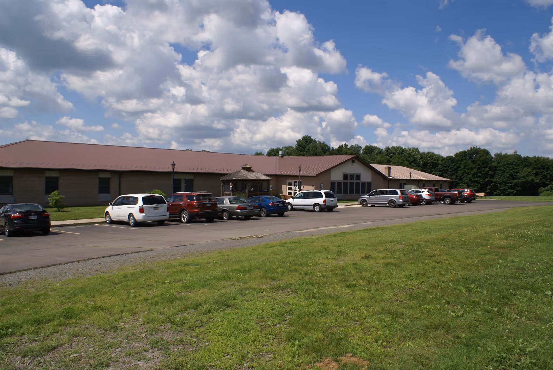 Exterior view of a single-story building with a brown roof and several parked cars in front under a partly cloudy sky. There is a small gazebo near the entrance and a grassy area in the foreground.