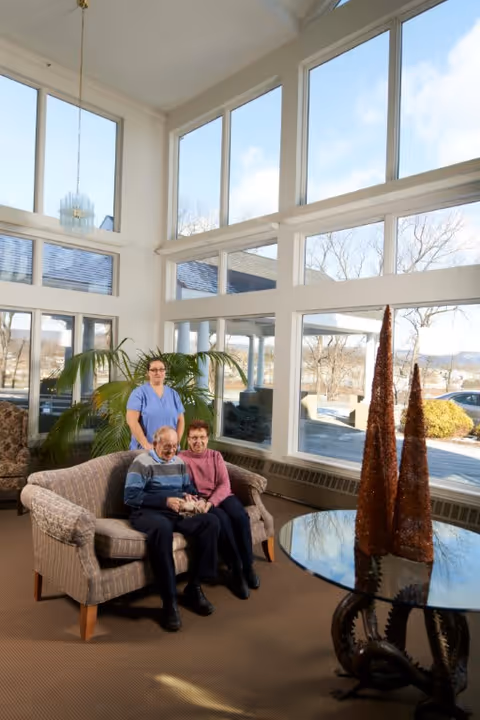 A bright and spacious living room area with large windows letting in natural light. An elderly couple is seated on a patterned loveseat, holding hands and smiling. Behind them stands a caregiver in blue scrubs. The room features a round glass table with decorative cone-shaped objects and a large green plant near the windows.