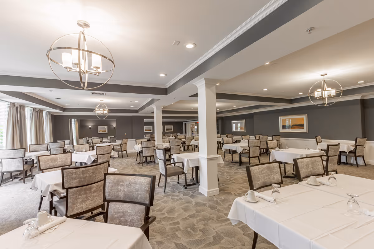 A spacious dining room with multiple tables covered in white tablecloths, each set with cups, napkins, and upside-down glasses. The room features beige upholstered chairs, gray walls with white wainscoting, modern circular chandeliers, and large windows with curtains allowing natural light.
