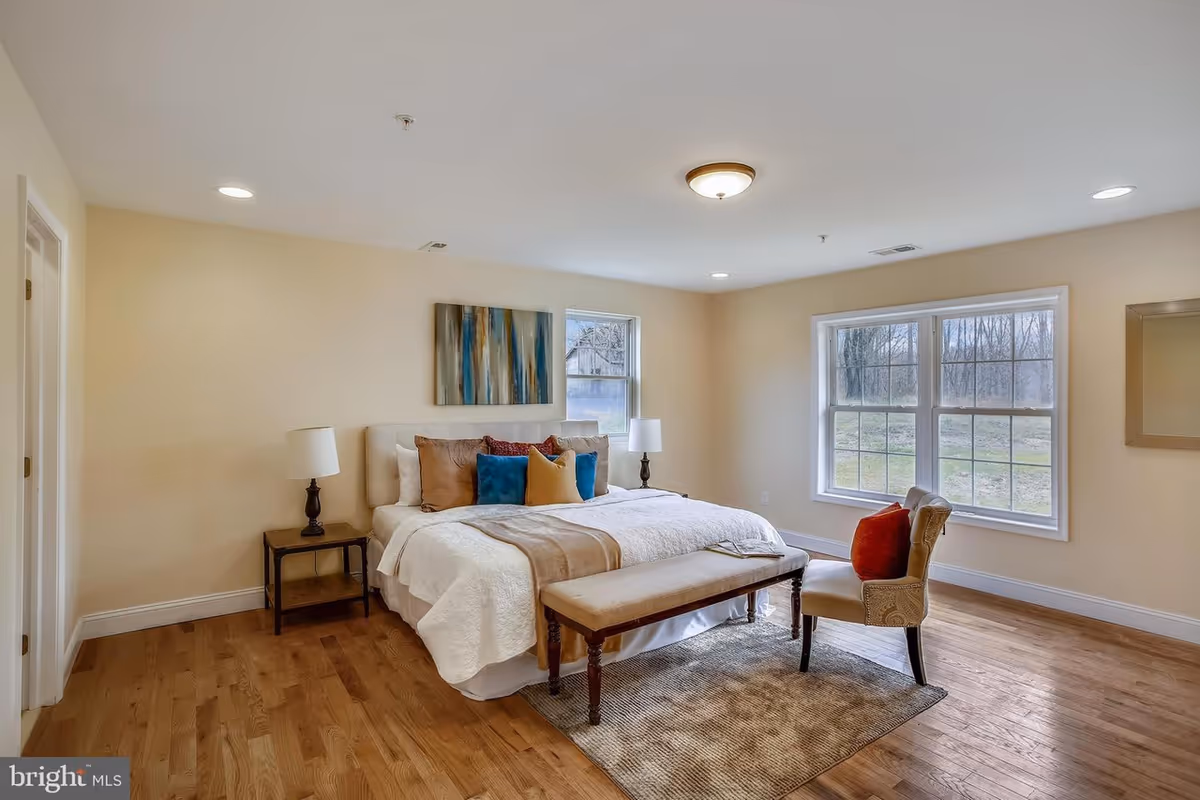 A bright bedroom with a large bed featuring white and beige bedding and multiple colorful pillows. There is a bench at the foot of the bed, a side table with a lamp, a cushioned chair with an orange pillow, two windows letting in natural light, a piece of abstract wall art above the bed, and hardwood flooring with a small area rug.