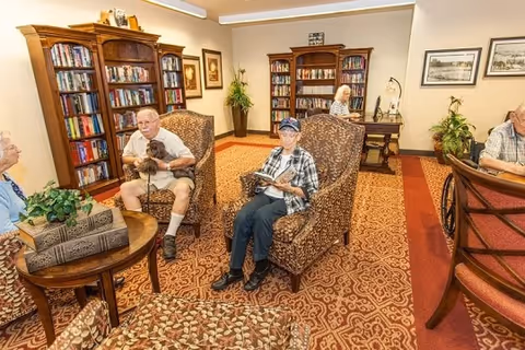 A cozy reading room in an assisted living facility with several elderly residents seated in patterned armchairs. The room features bookshelves filled with books, framed artwork on the walls, a patterned carpet, and a small table with decorative items and plants.