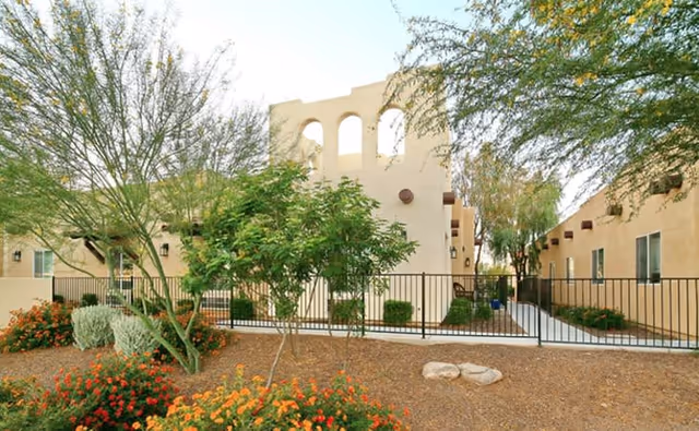 Outdoor view of a senior living facility with beige stucco buildings, desert landscaping including trees and flowering bushes, and a black metal fence enclosing a walkway between the buildings under a clear sky.