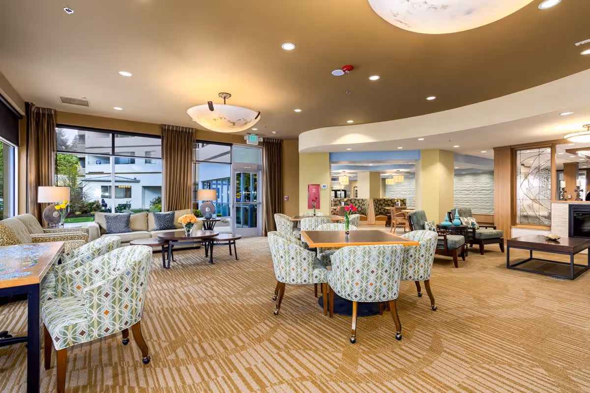 Bright, spacious common area with patterned dining chairs around tables, sofas, and large windows in a senior living facility.