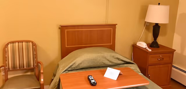 A small bedroom with a single bed covered with a green blanket, a wooden headboard, a wooden bedside table with a lamp and a telephone, a wooden chair with striped upholstery, and a wooden overbed table holding a TV remote and a folded card.