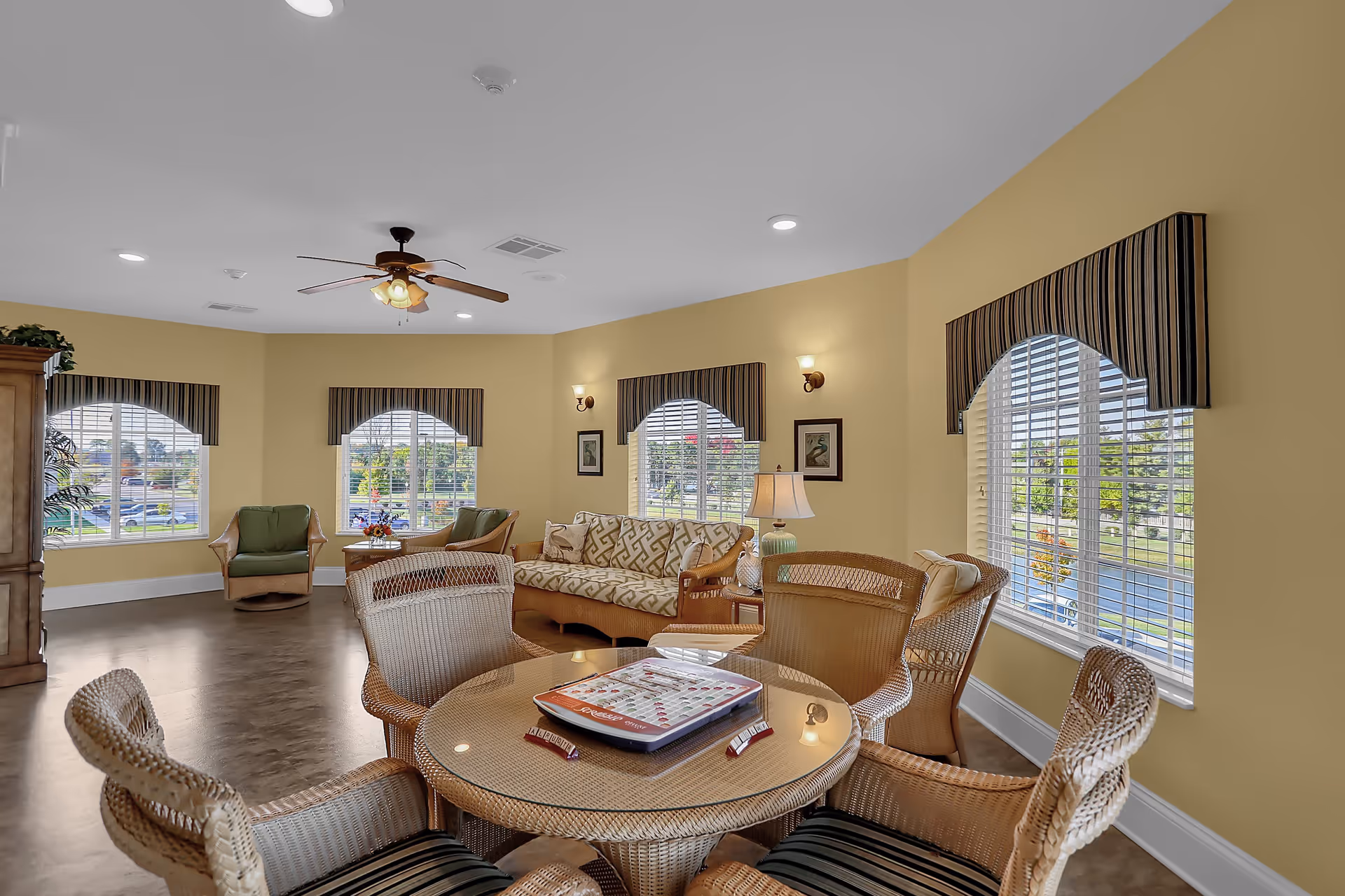 A cozy living room area with yellow walls and multiple windows with striped valances. The room features a round wicker table with four wicker chairs around it, a patterned sofa, two green armchairs, a wooden cabinet, and a ceiling fan with lights. There are framed pictures on the walls and a table lamp on a side table next to the sofa.
