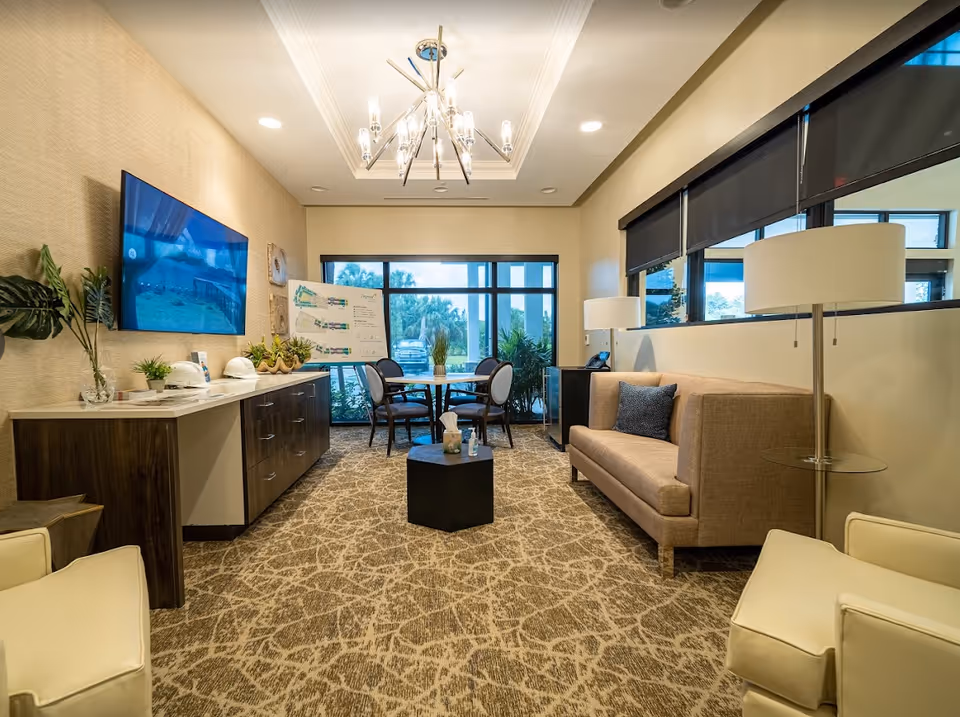 A modern, well-lit lounge area with a beige sofa, two white armchairs, a small black coffee table, and a round table with four chairs near large windows. A flat-screen TV is mounted on the wall above a wooden cabinet with plants and hard hats. The room has patterned carpet flooring, a chandelier, and large windows with black roller shades.