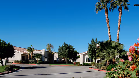View of a residential-style senior living facility with single-story buildings surrounded by palm trees, bushes, and flowering plants under a clear blue sky.