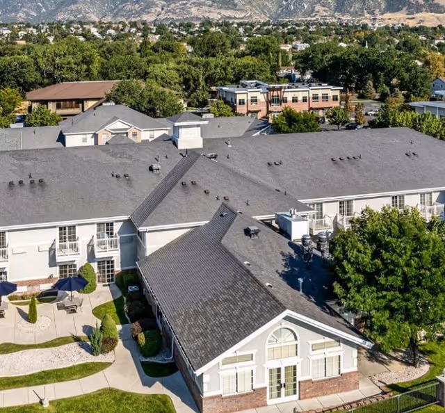 Aerial view of a senior living complex with connected buildings, landscaped courtyard, walkways, and surrounding trees and distant mountains.