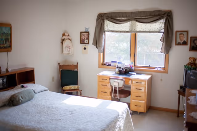 A cozy bedroom with a bed covered in a white quilt, a wooden chair with a cushion, a wooden desk with drawers under a window with blinds and curtains, and a small TV on a stand with a teddy bear on top. The walls have framed pictures and a decorative doll hanging.