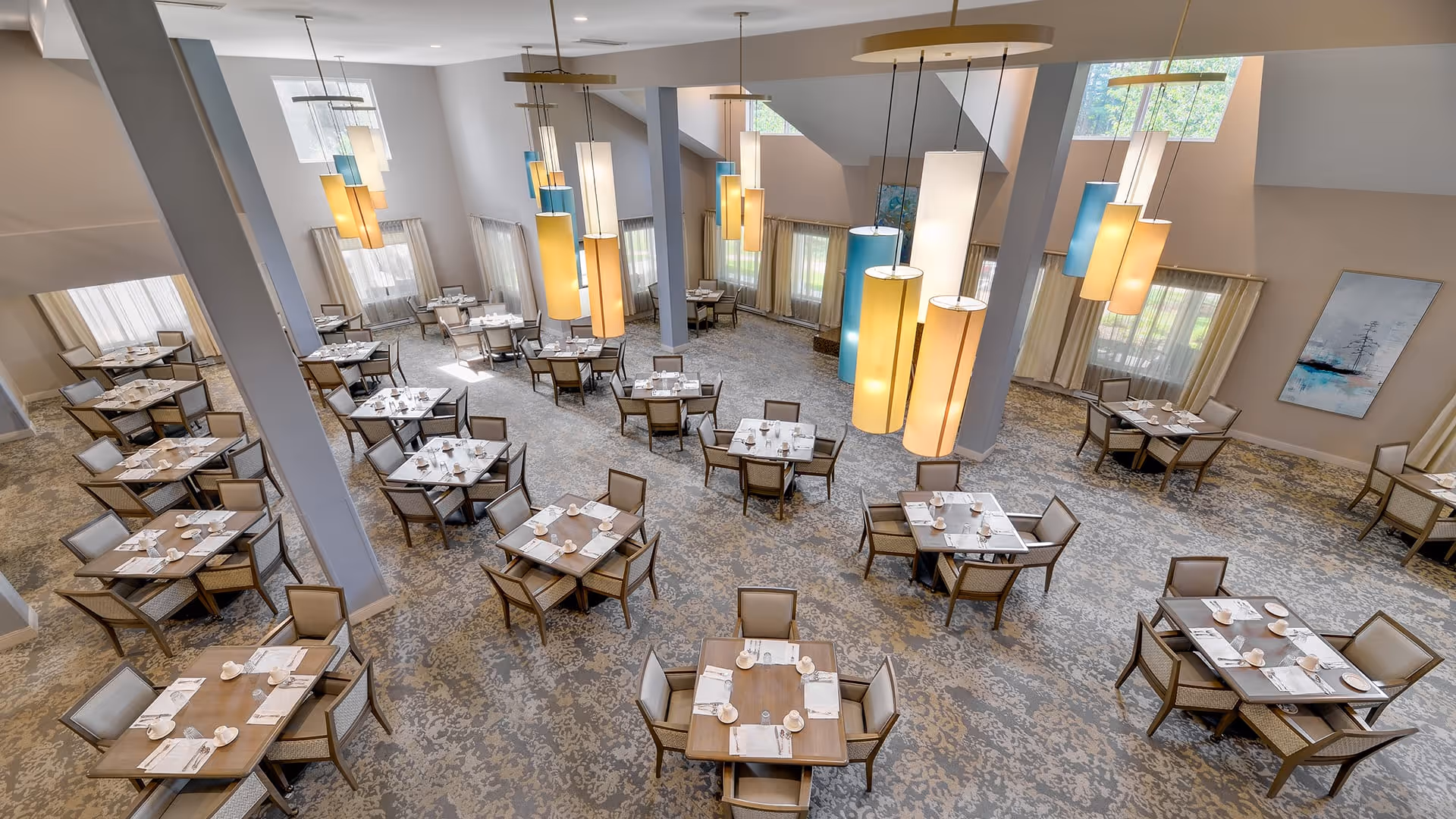 Spacious dining room with multiple square tables each set with white napkins, cups, and plates. The room features large windows with curtains, modern hanging pendant lights in various colors, and a patterned carpet floor. The walls are light-colored with some artwork displayed.