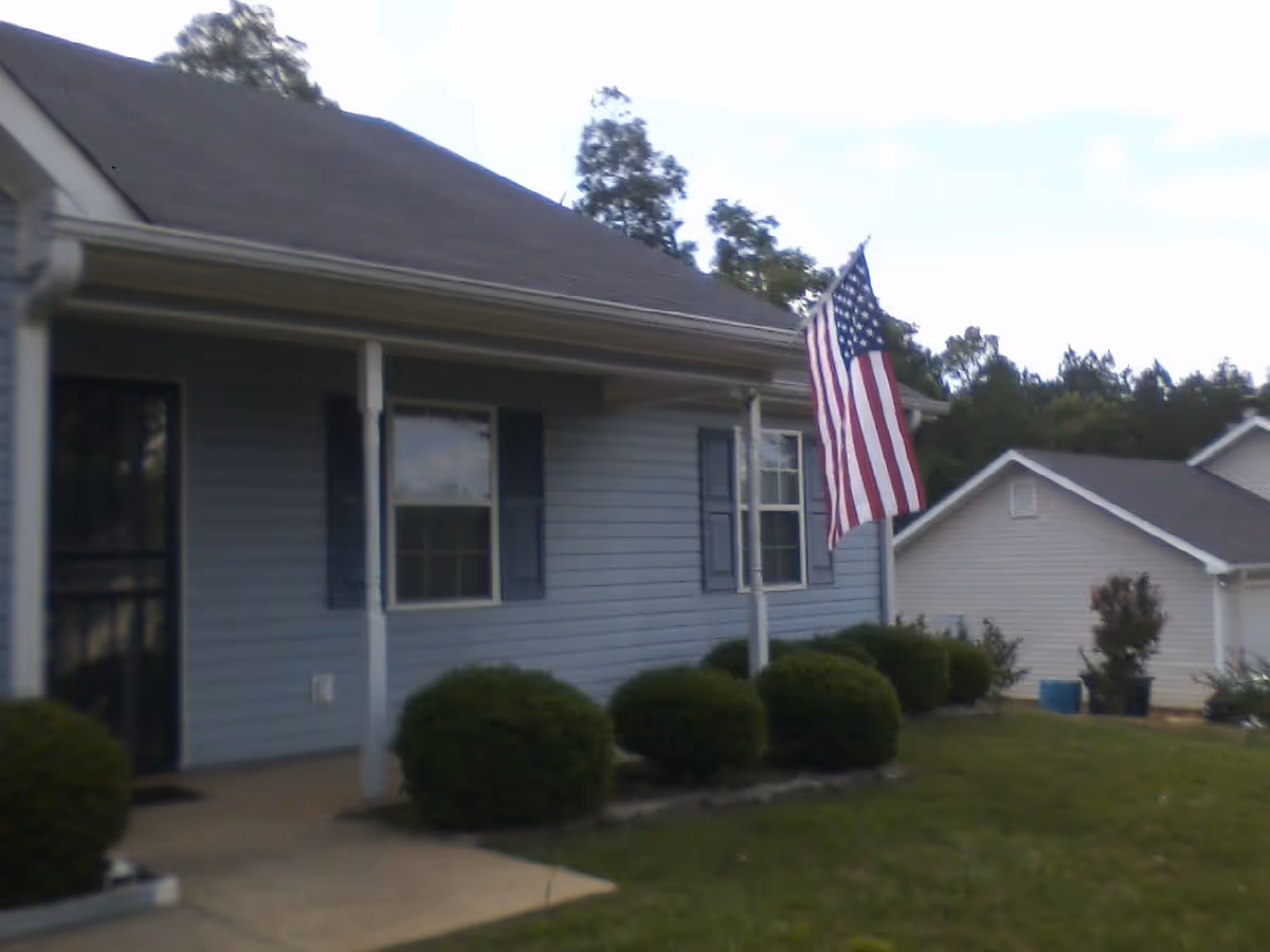 Front exterior of a single-story house with a porch, American flag, and trimmed shrubs.