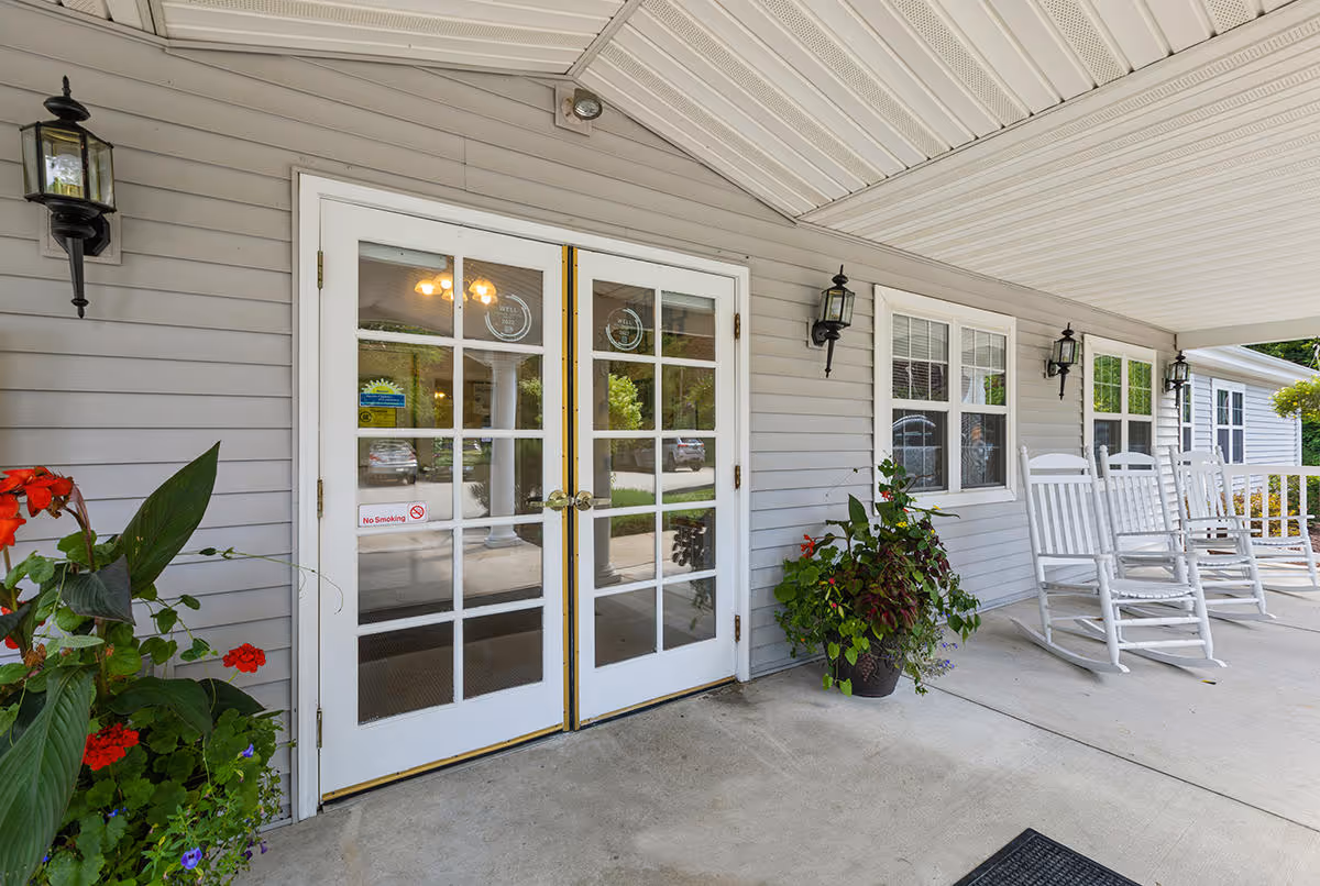 Covered porch area at the entrance of The Addison of Uniontown facility featuring double glass doors, potted plants with red and green foliage, white rocking chairs, and wall-mounted lantern-style lights.