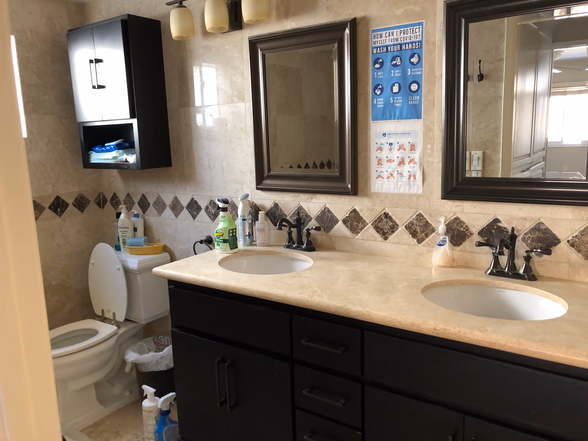A bathroom with a double sink vanity featuring dark cabinets and beige countertops. Above the sinks are two framed mirrors and a wall-mounted light fixture with three lights. The wall is tiled with beige tiles and a decorative diamond pattern. To the left is a toilet with the lid open, a wall-mounted cabinet above it, and various cleaning supplies on the countertop and floor. There are two posters on the wall with COVID-19 handwashing instructions.