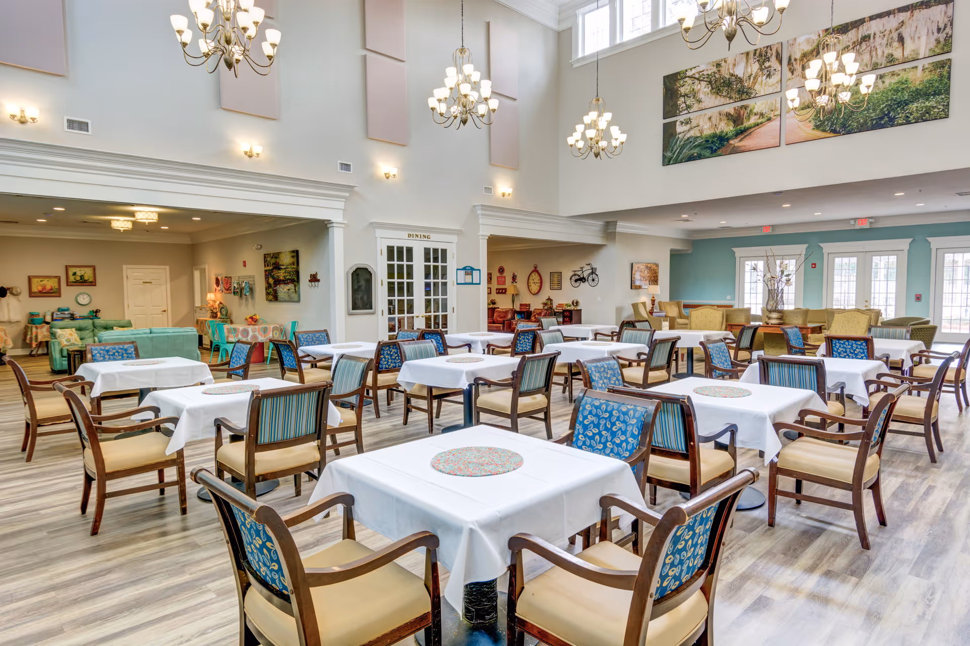 A spacious dining room with multiple tables covered in white tablecloths and surrounded by wooden chairs with patterned cushions. The room features high ceilings with chandeliers, large windows, and decorative wall art. There is a seating area with couches and armchairs in the background.