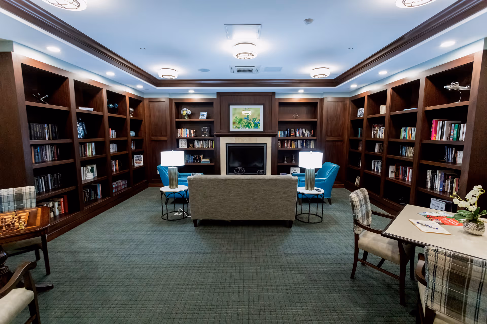 A cozy library room with dark wooden bookshelves lining the walls filled with books and decorative items. In the center, there is a beige sofa facing a fireplace with a framed picture above it. Two blue armchairs flank the sofa, each with a side table and lamp. On the right side, there is a table with two plaid upholstered chairs and a small plant on top. The room has a green carpet and recessed lighting in the ceiling.