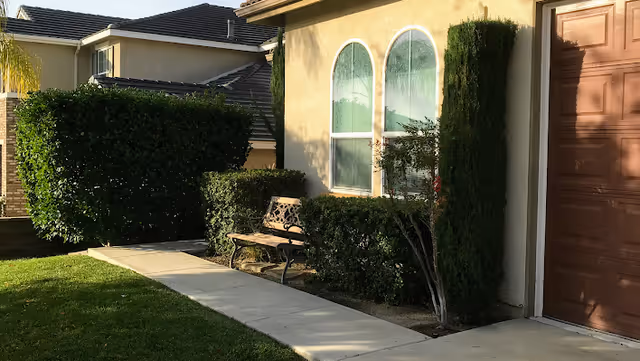 Outdoor walkway alongside a beige building with two arched windows and a brown door. There is a metal bench surrounded by trimmed bushes and a small tree, with a green lawn and another building visible in the background.